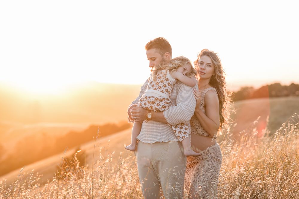 Expectant mother, father, and young daughter embrace at sunset in a golden sunlit meadow. Lakes Region maternity family photographer