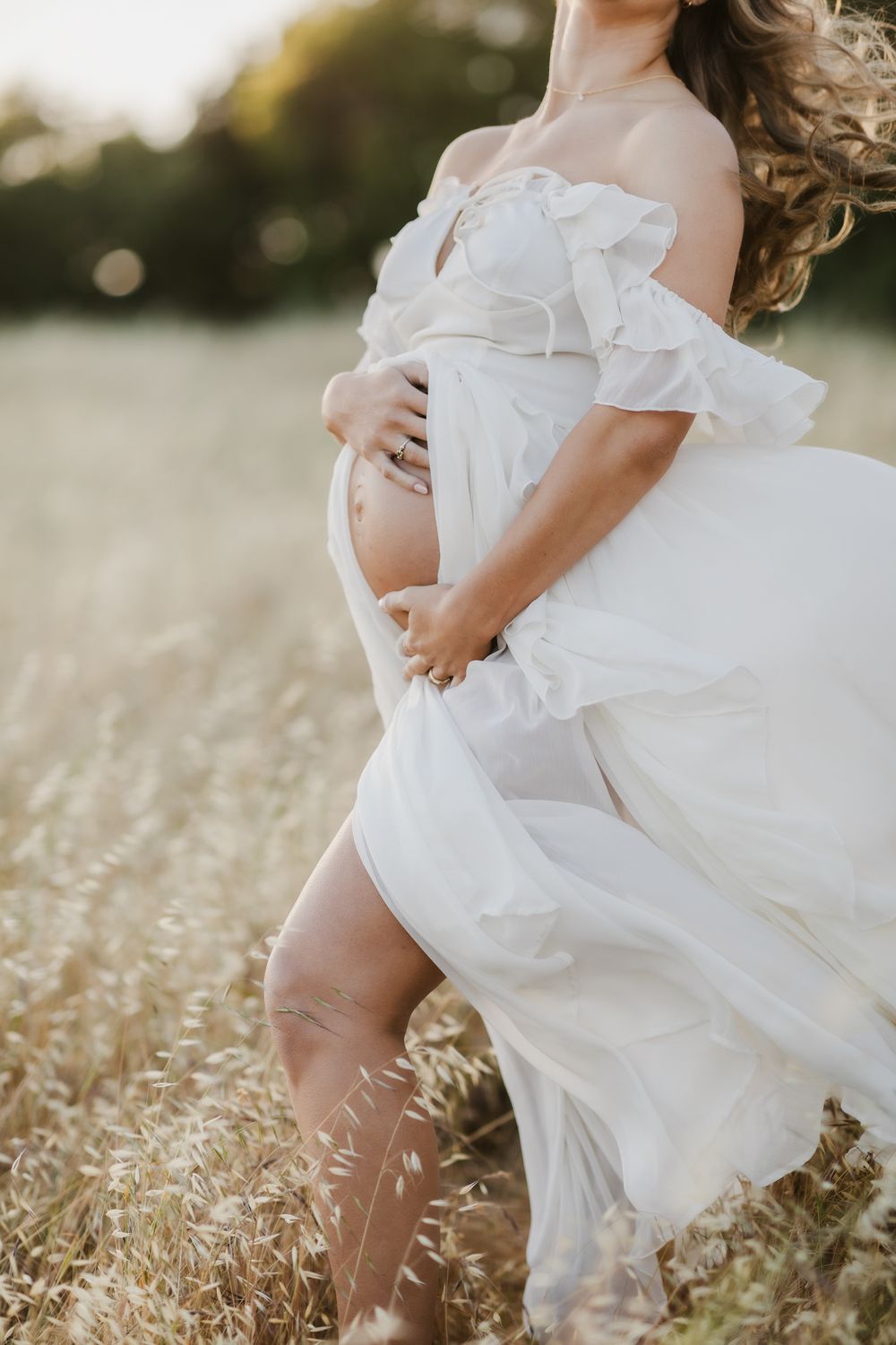 Expectant mother in white gown holds baby bump in golden field as hair blows in wind. Harvard Hollis Rye maternity photographer