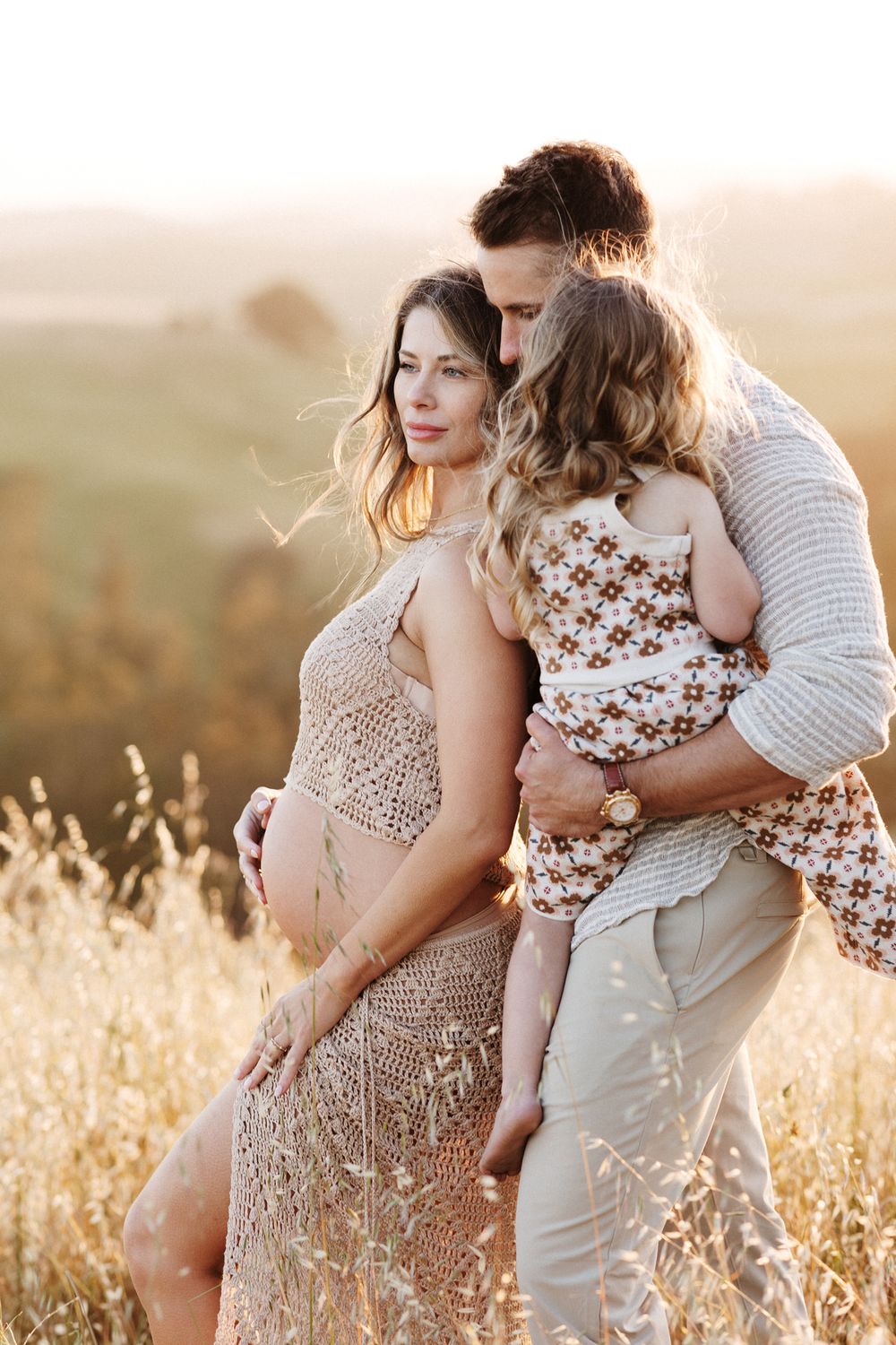 Husband holds young daughter and embraces expectant mother wearing gold crochet outfit in golden wheat field. Groton maternity photographer