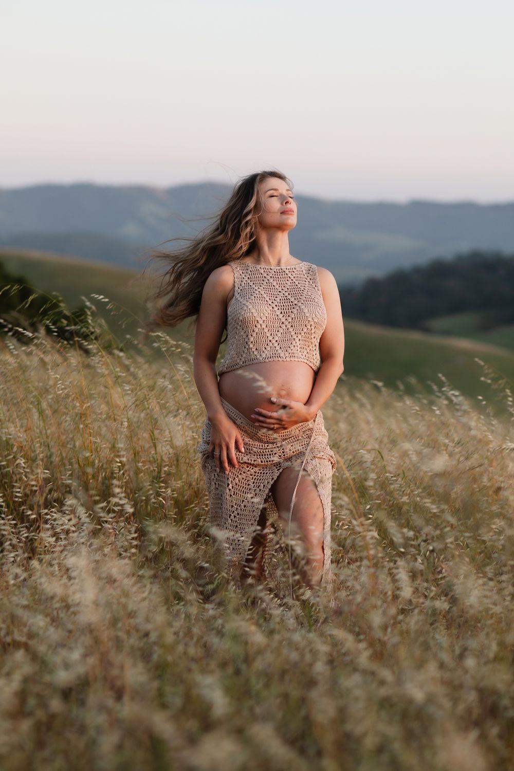 Expectant mother wearing tan crochet outfit tilts head back and poses in grassy field at blue hour. Concord York maternity photographer