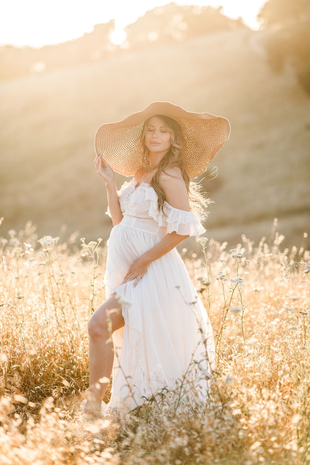 Expectant mother in white gown and wide-brim straw hat poses in golden sunlit field at golden hour. Seacoast Hampton maternity photographer