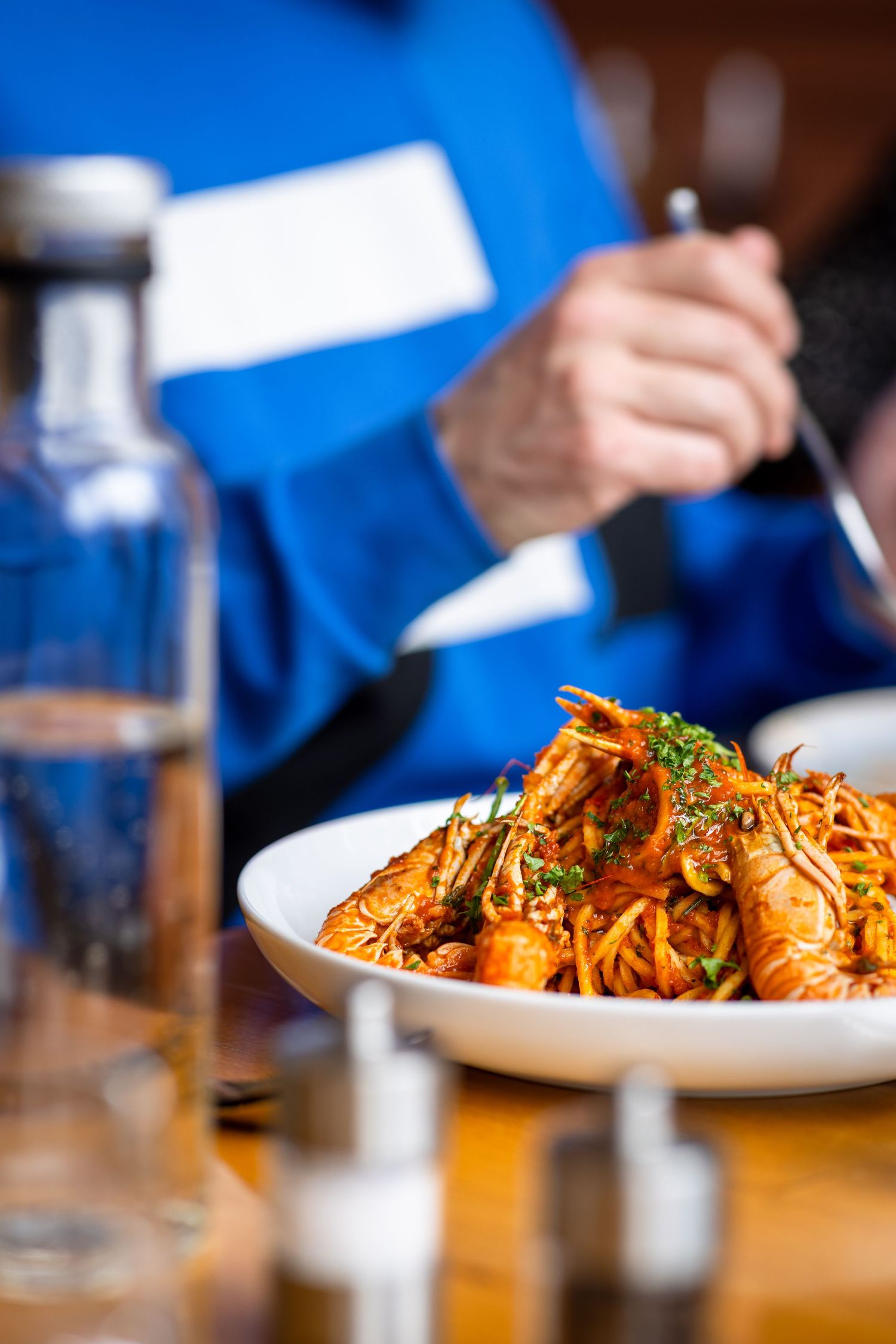A white bowl of seafood pasta with shrimp in tomato sauce, served at a restaurant table with a person in blue dining.