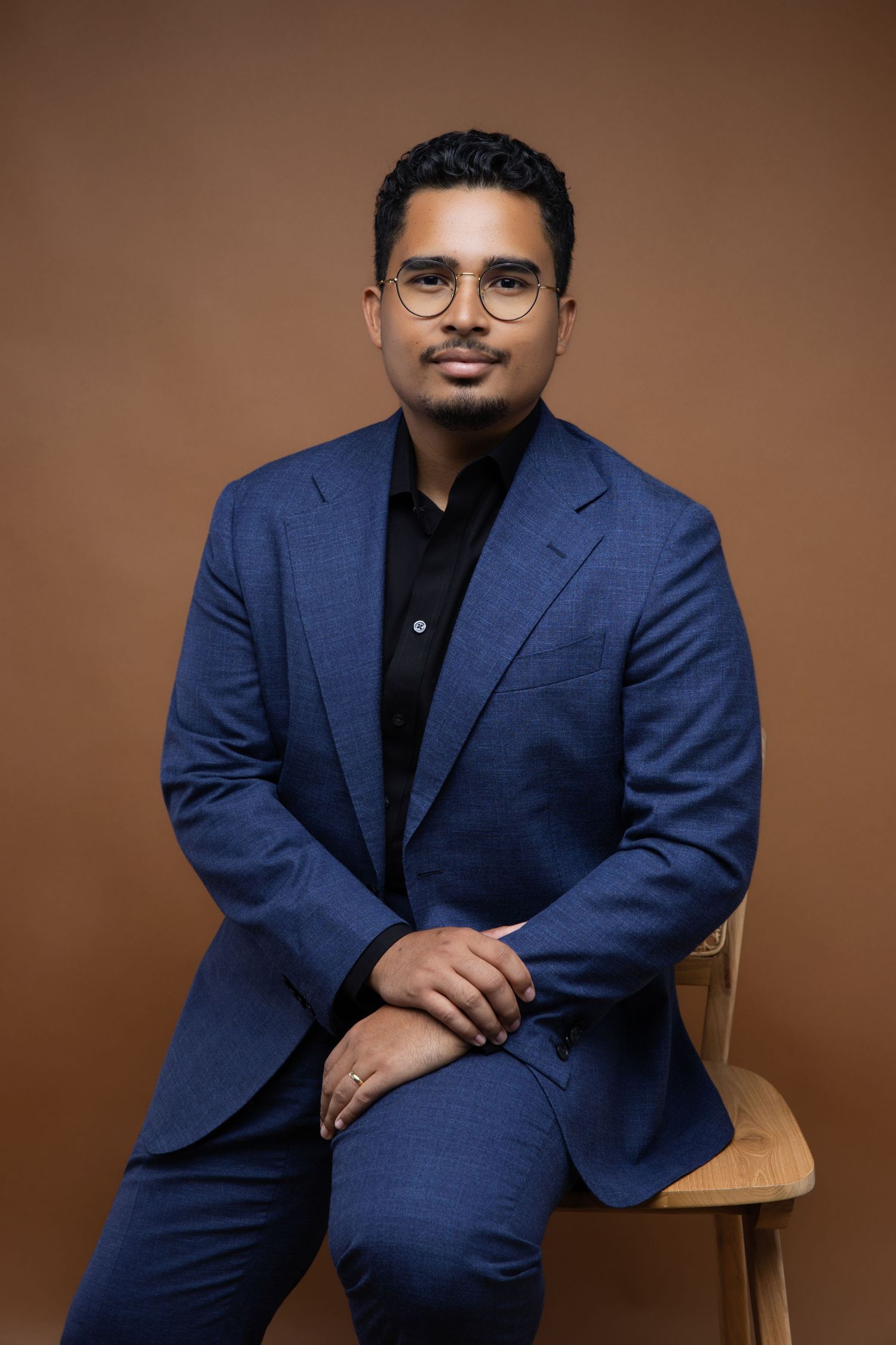 Professional studio portrait of someone in a navy blue blazer and black shirt against a brown backdrop.