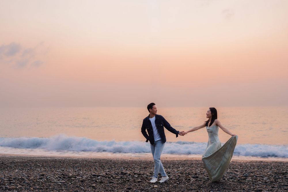 Couple holding hands walking on a pebble beach at sunset with gentle ocean waves in the background.