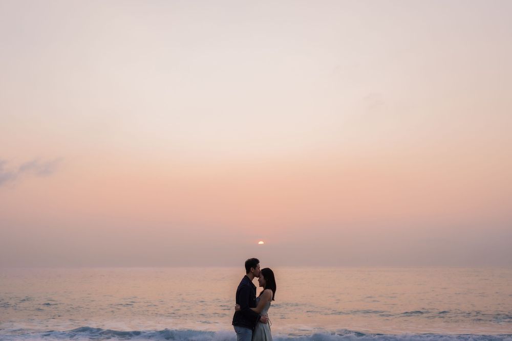 Couple sharing a romantic kiss on the beach at sunset with soft pink and orange hues reflecting over the ocean.