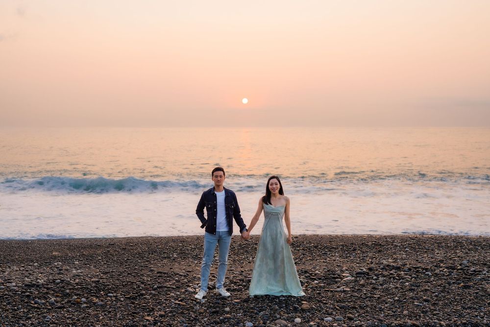 Couple holding hands on rocky beach at sunset with ocean waves and pink sky backdrop.