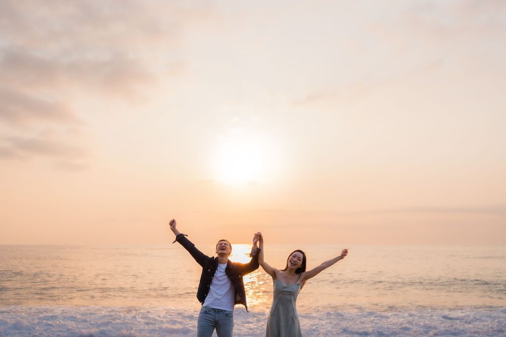 Couple raising arms joyfully on a beach at golden sunset with ocean waves in the background.