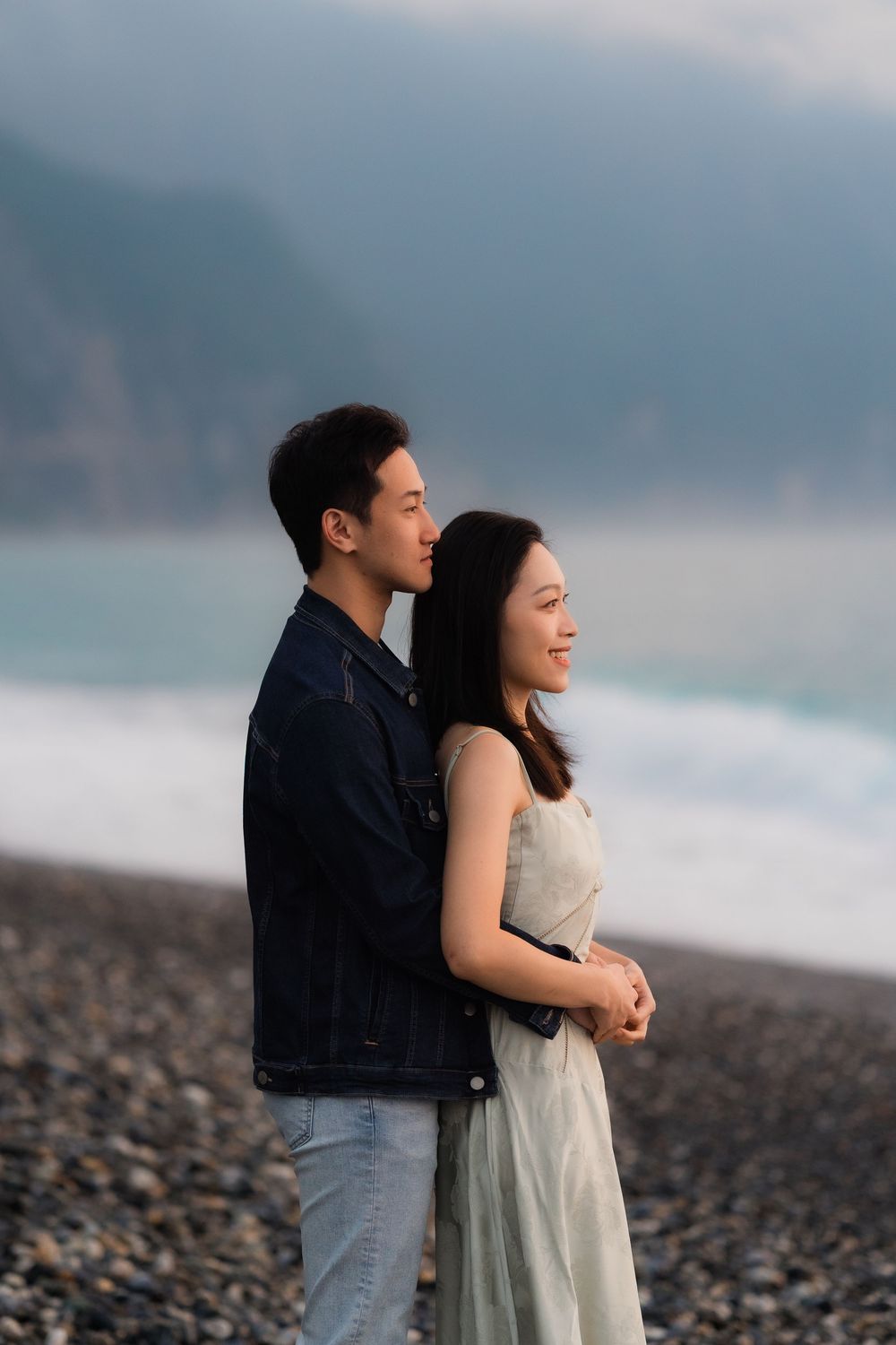 A young Asian couple embraces on a rocky beach, gazing toward the ocean with misty mountains in the background.