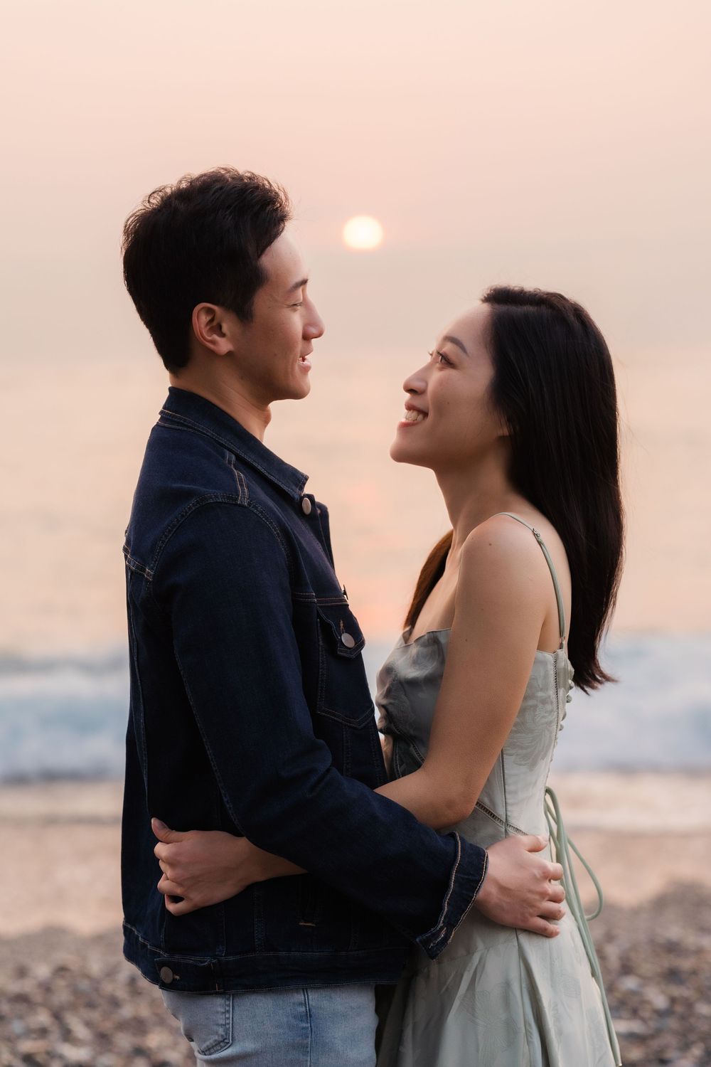 A smiling Asian couple embracing on a pebble beach at sunset, sharing a romantic moment by the ocean.