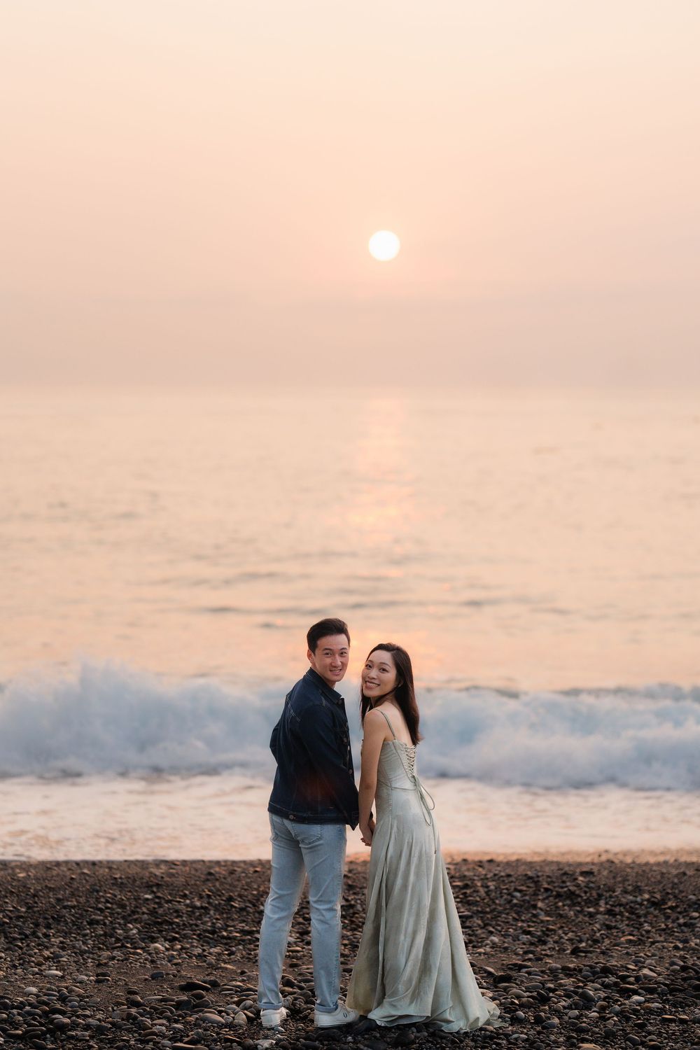Couple holding hands on a rocky beach at sunset with golden ocean reflections behind them.