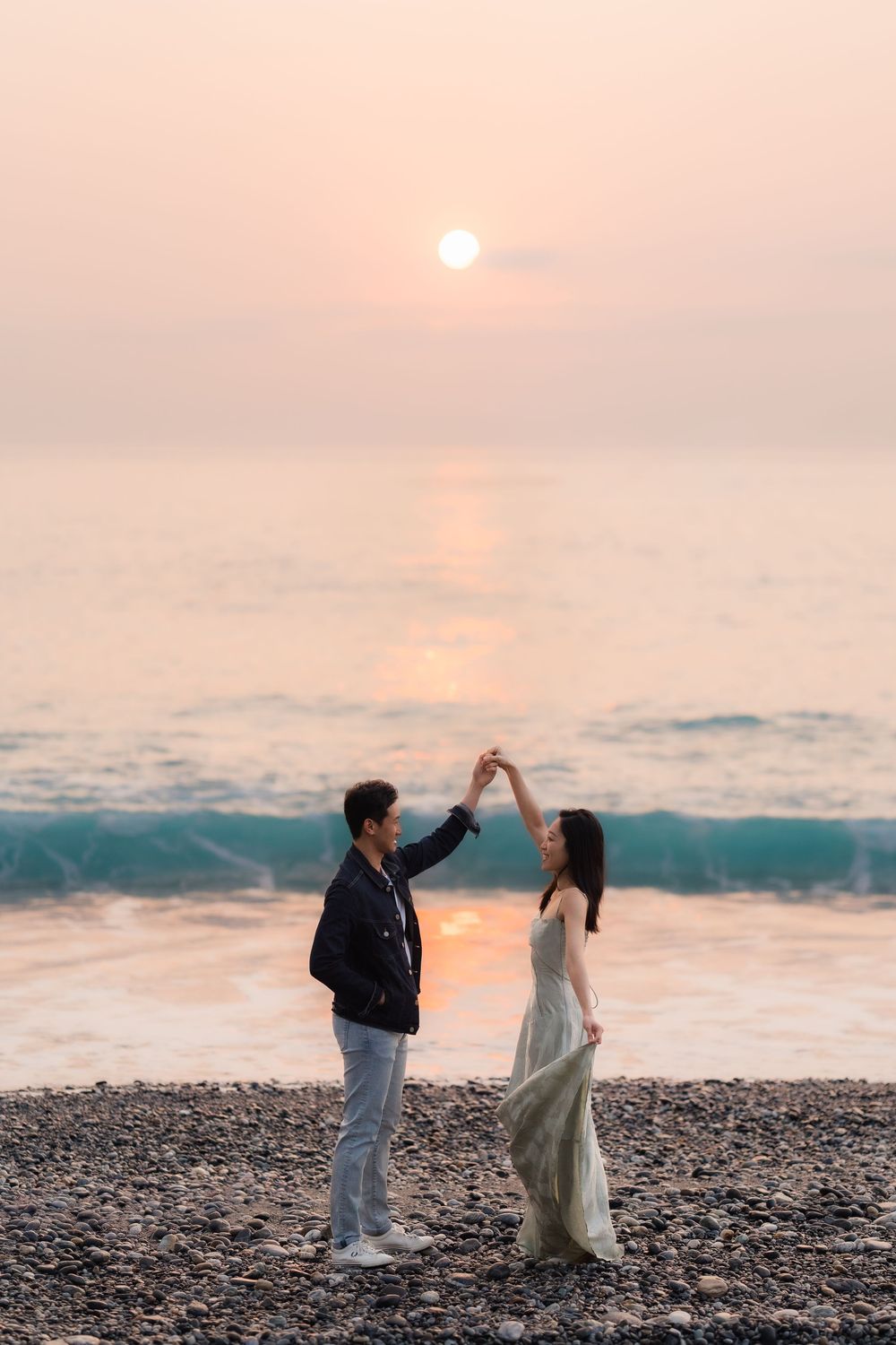 Couple dancing on rocky beach at sunset, waves and glowing sun reflecting on calm ocean water behind them.