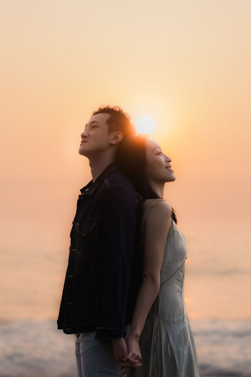 Couple holding hands back-to-back at sunset beach, golden sky glowing behind them.