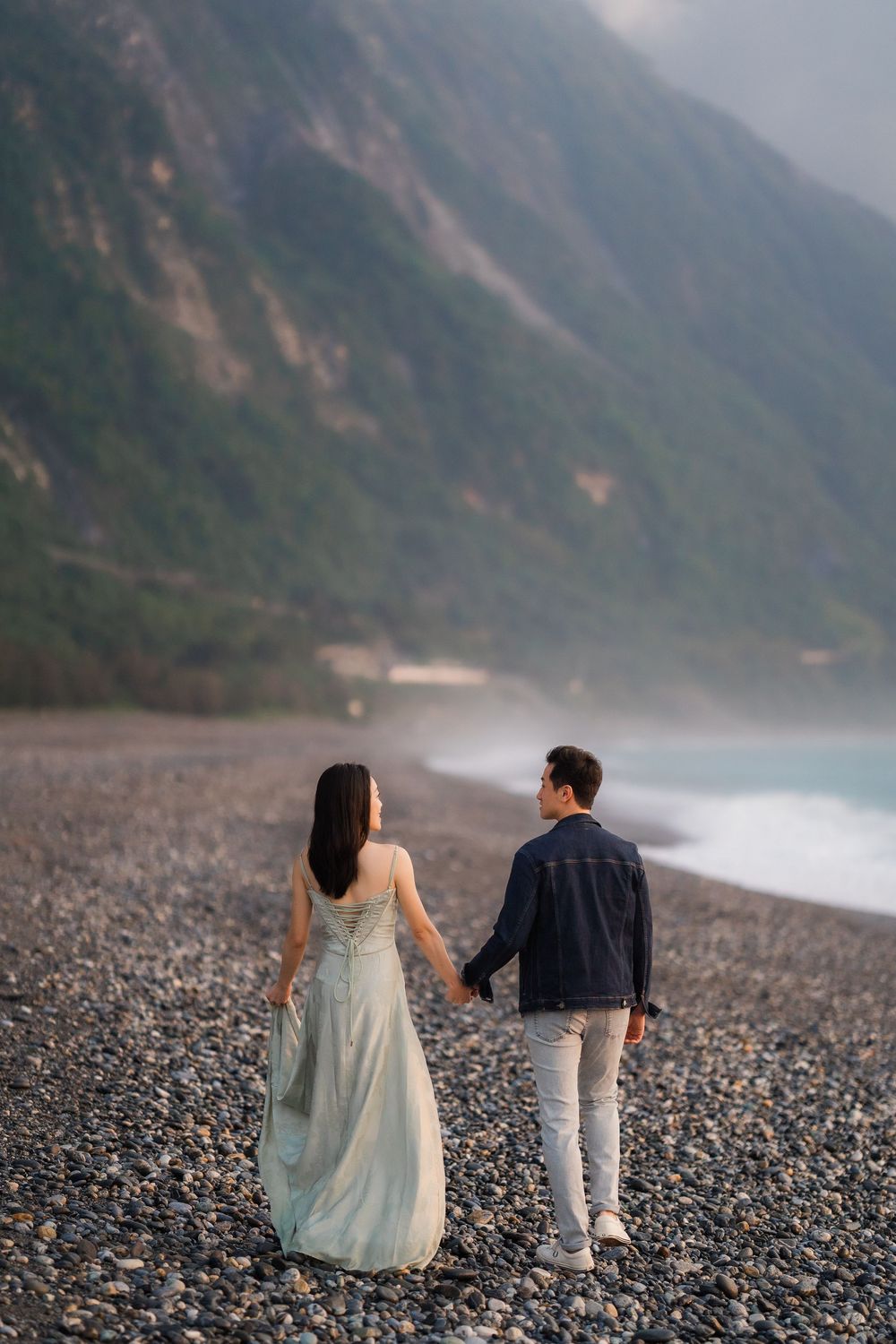 Couple holding hands walking on rocky beach with misty mountains and ocean waves in background.
