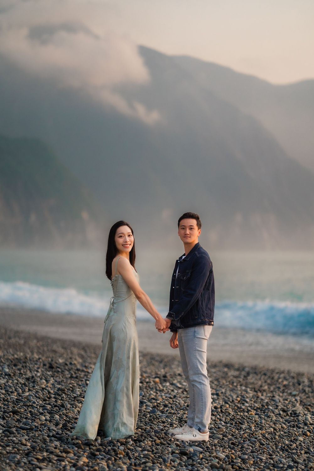 Couple holding hands on a rocky beach with dramatic misty mountains and ocean waves.