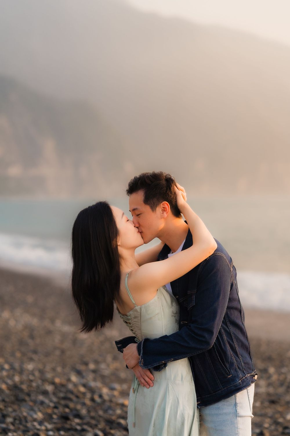 Couple sharing a romantic kiss on a pebble beach with misty mountains at sunset.