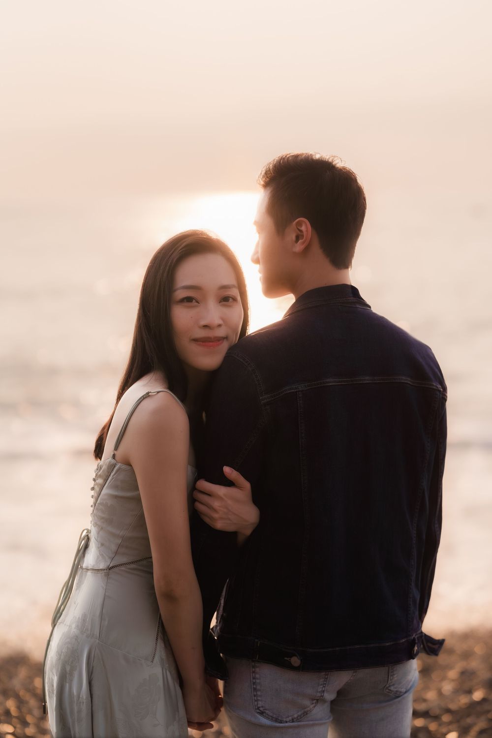 Romantic couple embracing at sunset on pebble beach, woman smiling at camera with golden light.