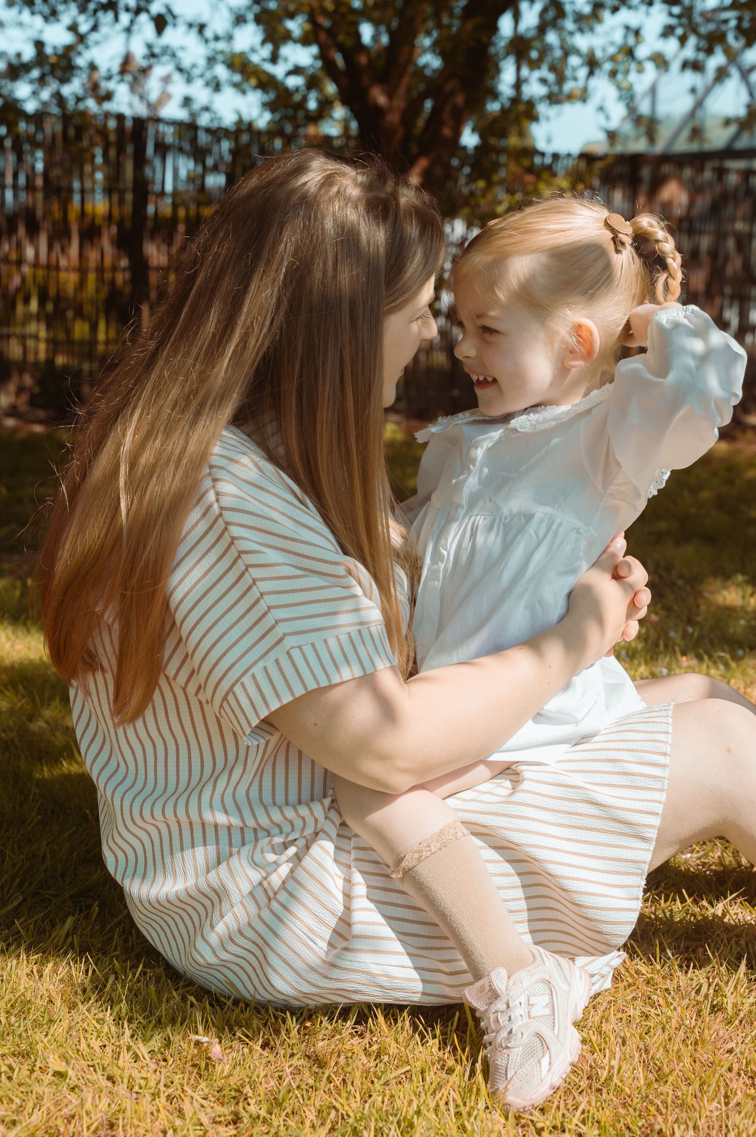 A mother sits on green grass holding her laughing toddler daughter, both wearing light dresses on a sunny day outdoors.