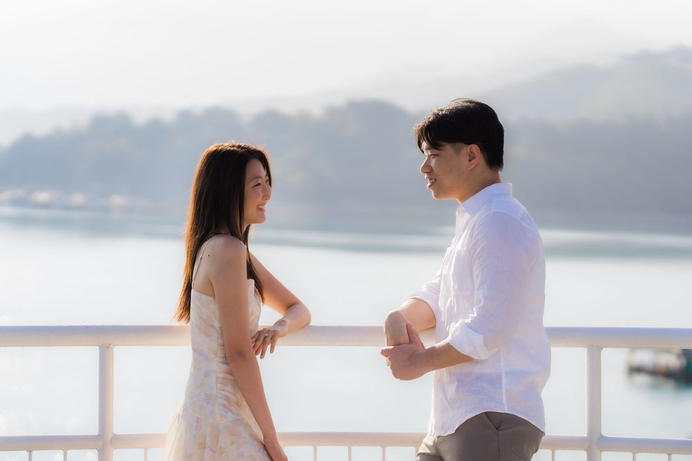 A young Asian couple shares a romantic moment on a waterfront balcony with scenic mountain views.