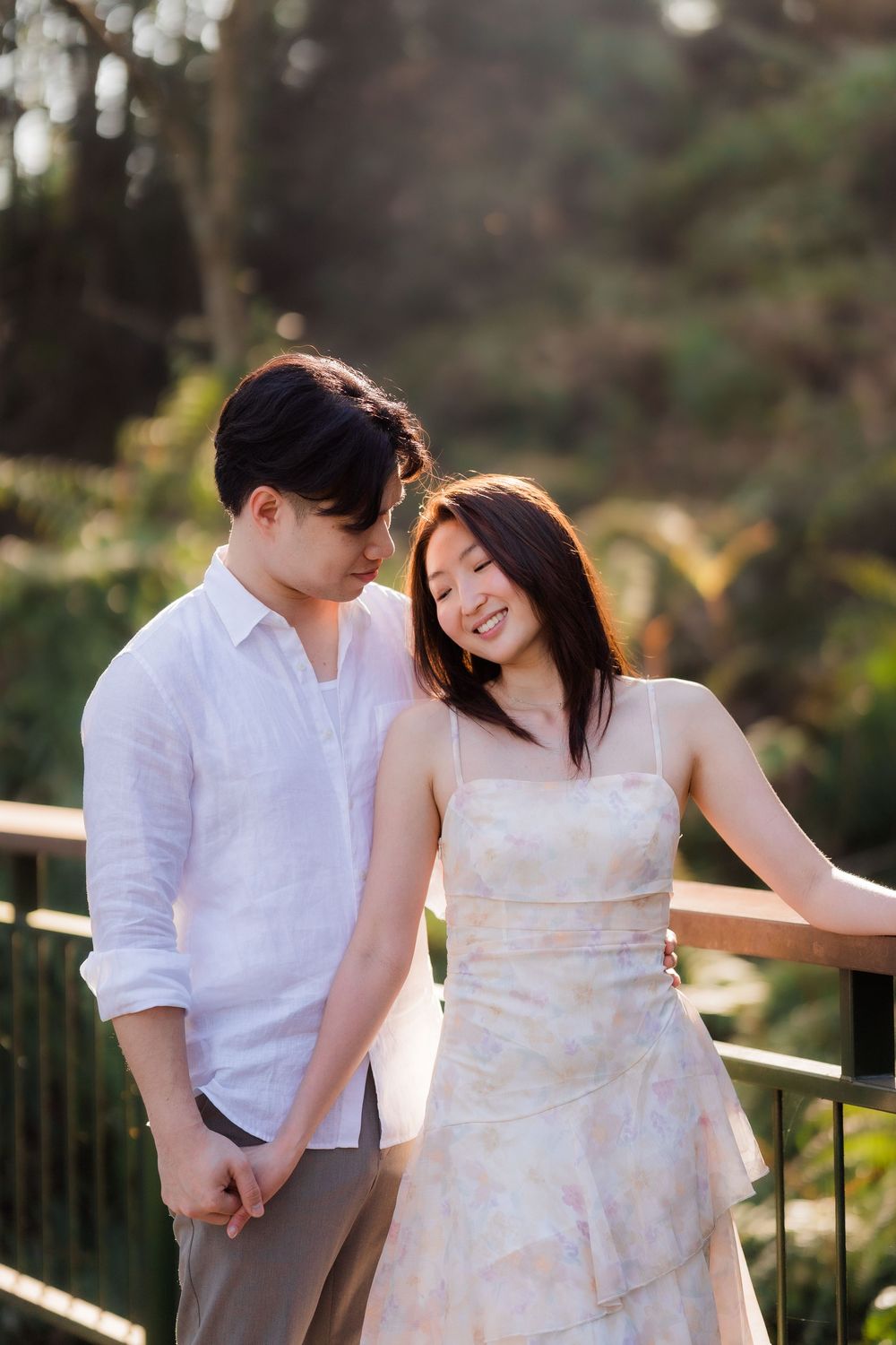 A young Asian couple shares a tender moment on a wooden bridge surrounded by lush greenery in warm sunlight.