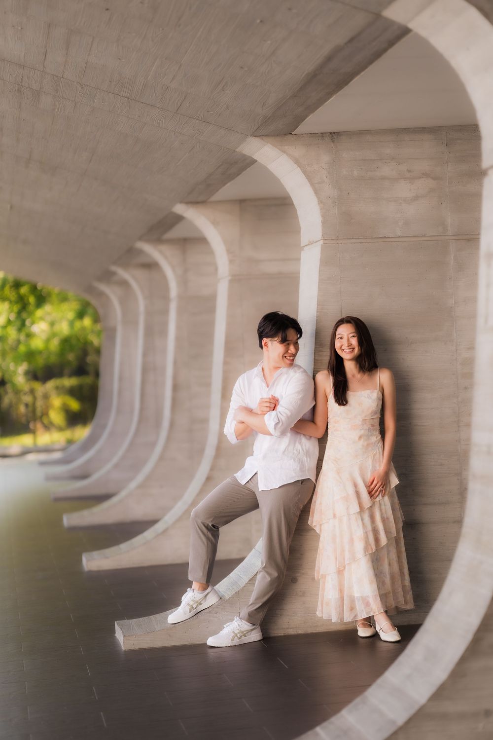 Couple posing under curved concrete arches, woman in floral dress and man in white shirt, smiling in modern architectural setting.