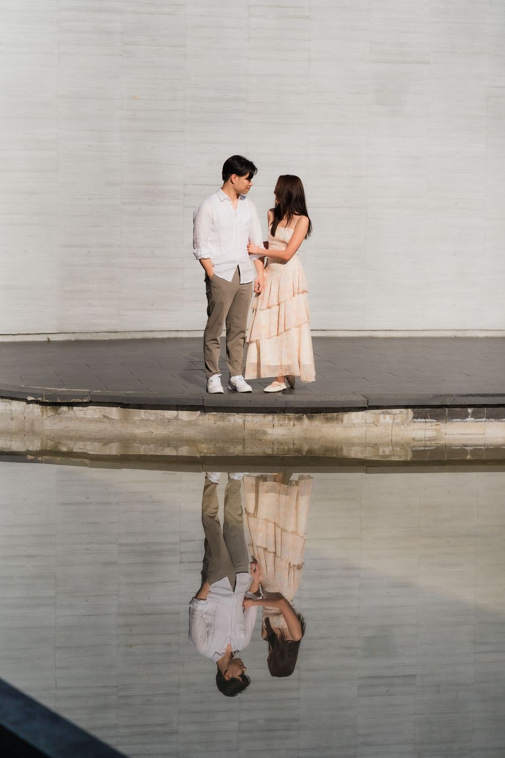 Couple in love standing by a reflective pool, man in white shirt and woman in pink dress, mirrored in water.