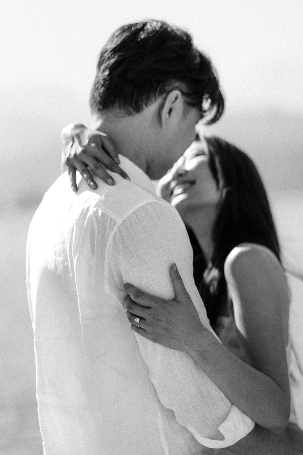 Black and white photo of a smiling couple embracing outdoors, woman wearing a ring, man in white linen shirt.