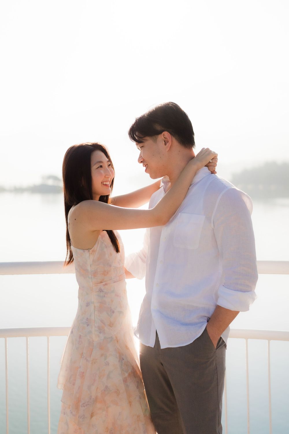 A couple shares a romantic moment on a waterfront balcony, the woman in a floral dress smiling at her partner.