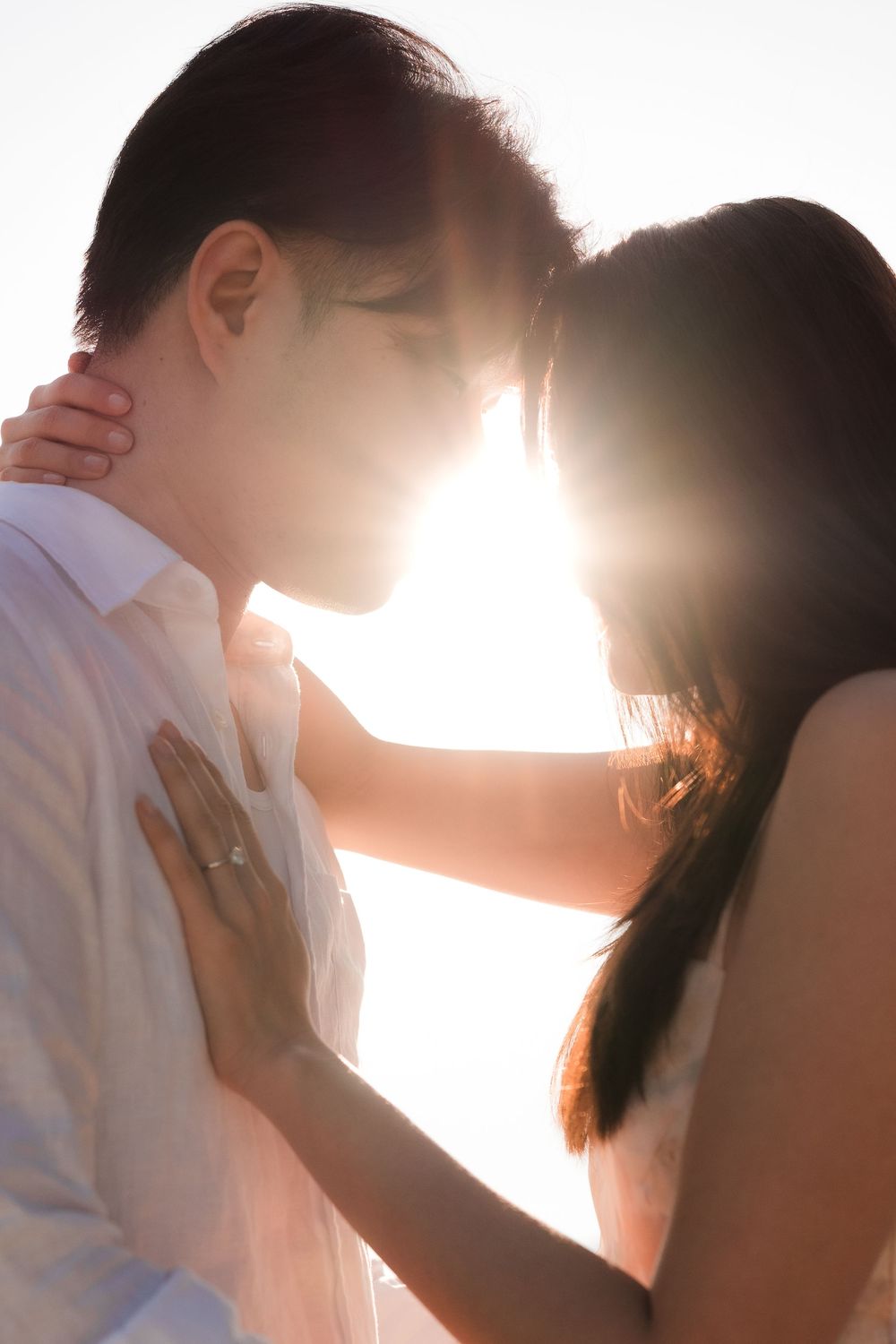 Couple sharing a romantic moment backlit by golden sunlight, woman wearing engagement ring resting hands on mans chest.
