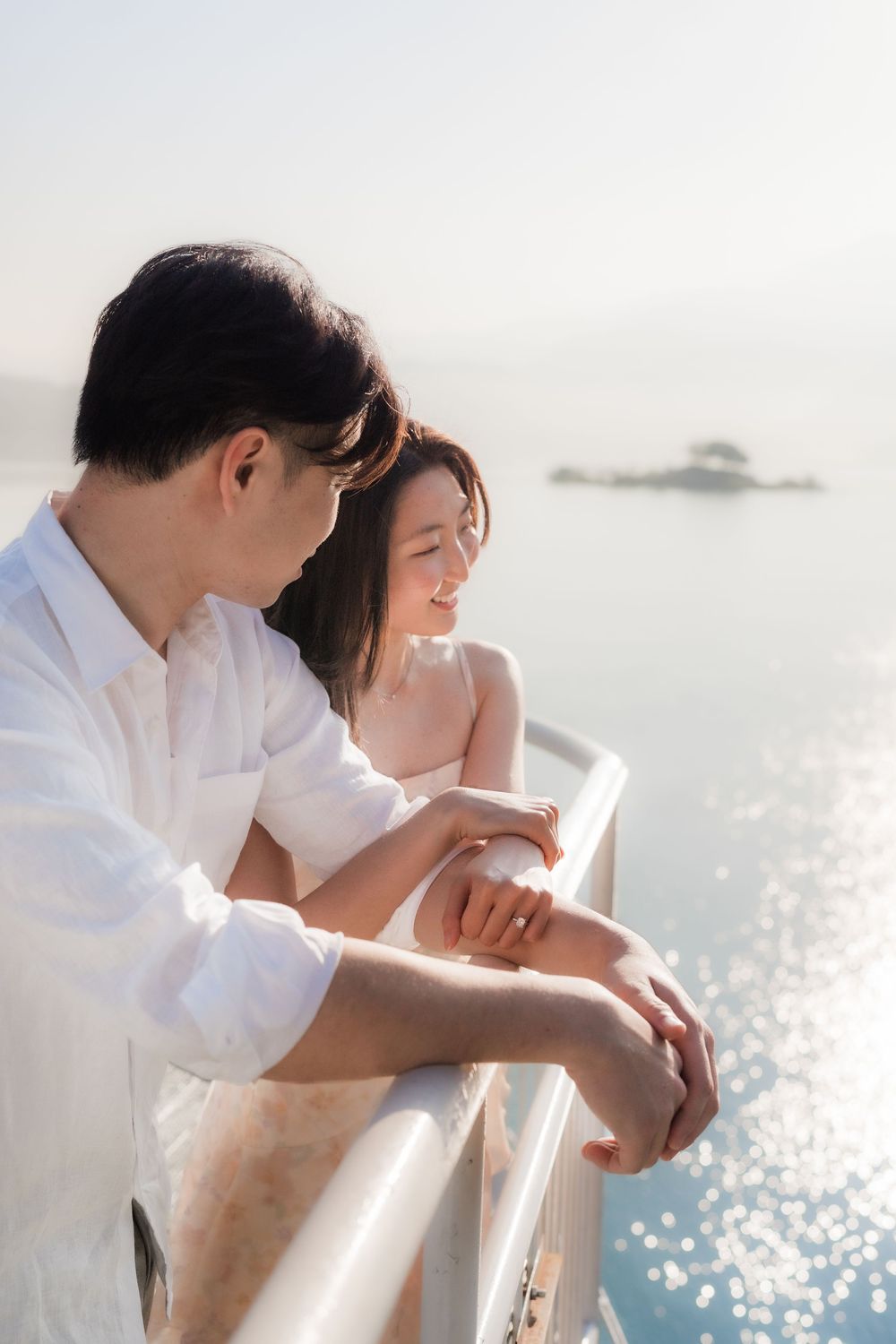 Romantic Asian couple leaning on white railing overlooking sparkling ocean, dressed in white outfits on a sunny day.