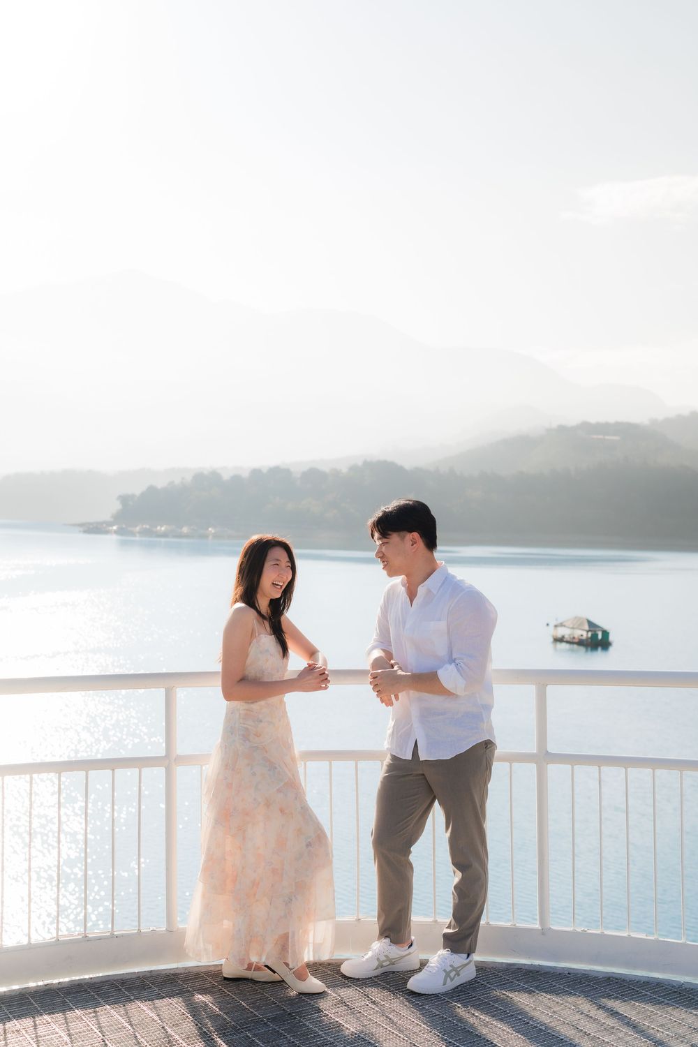 Couple holding hands on a scenic overlook with a serene lake and misty mountains in the background.
