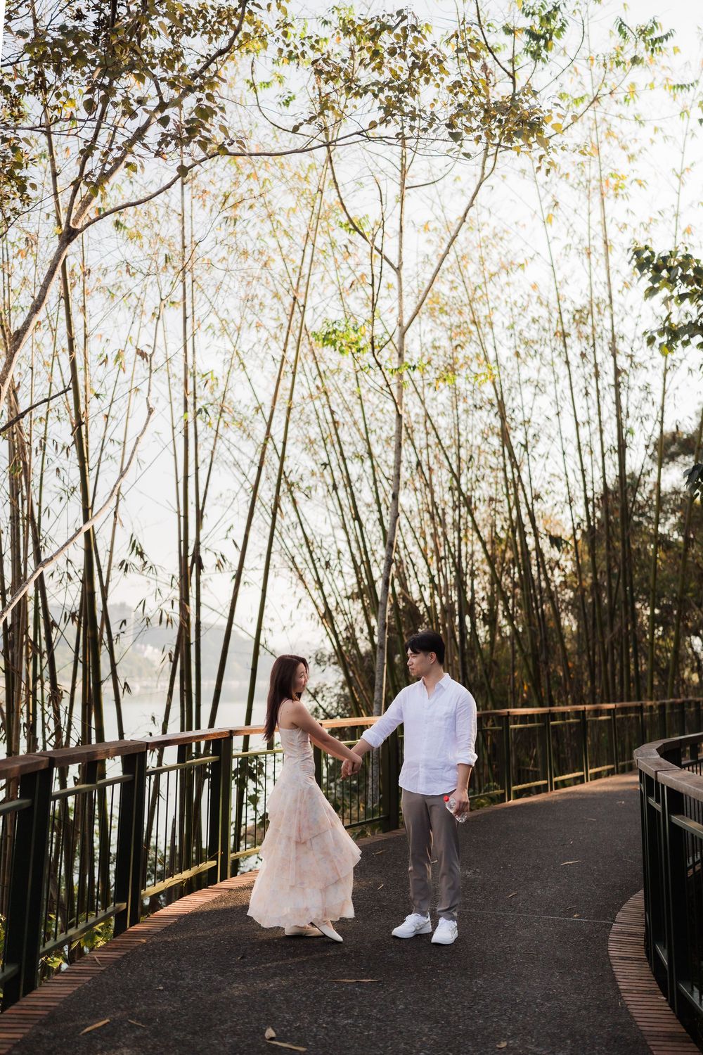 Asian couple holding hands on a wooden walkway surrounded by tall bamboo trees near a serene lake.