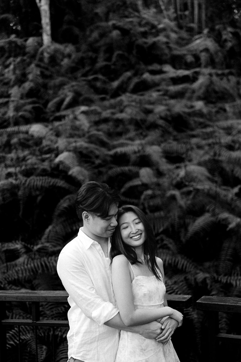 Black and white photo of a smiling couple embracing outdoors with lush fern foliage in the background.