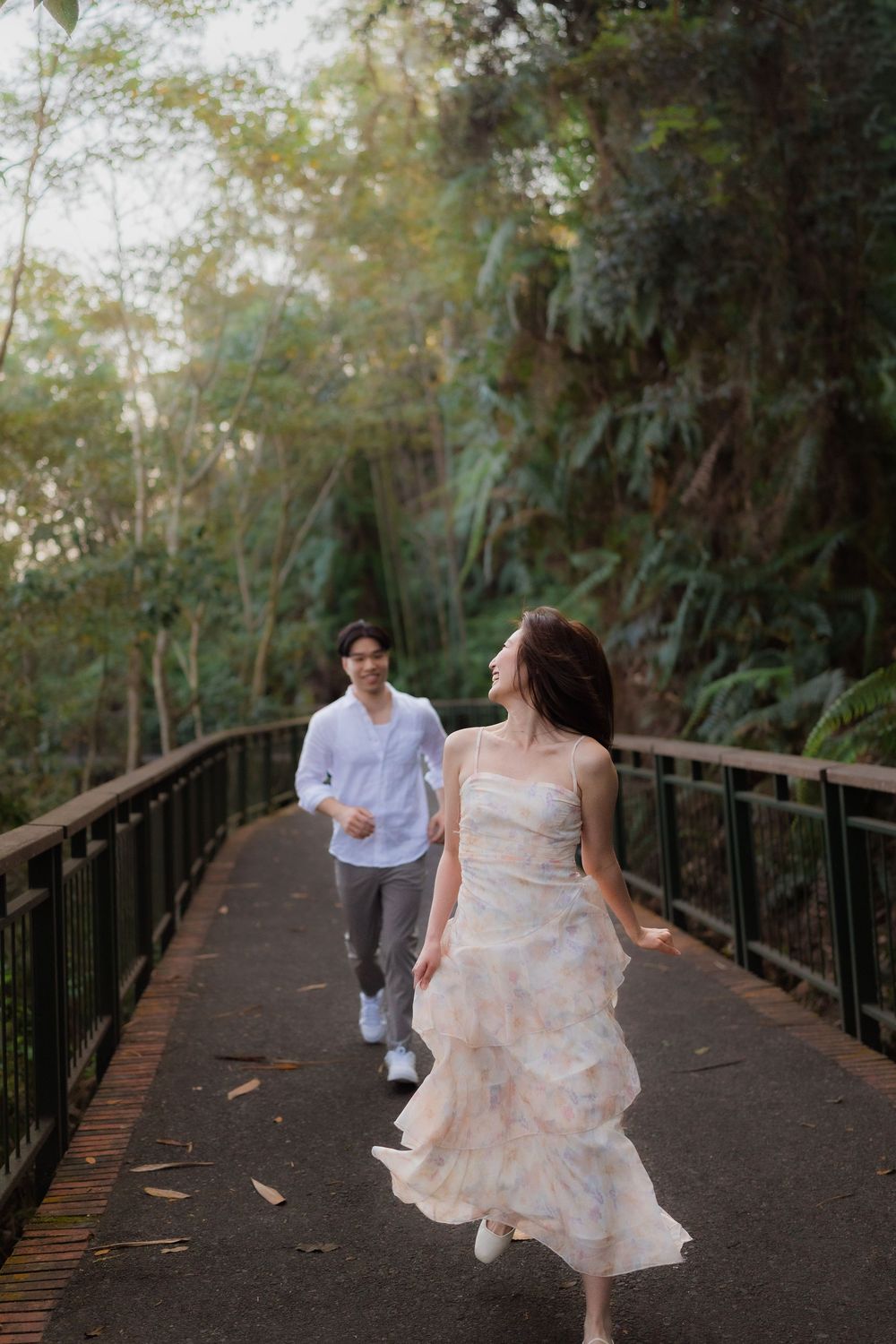 A couple runs along a lush forest bridge, woman in floral dress, man in white shirt, during a romantic outdoor photoshoot.
