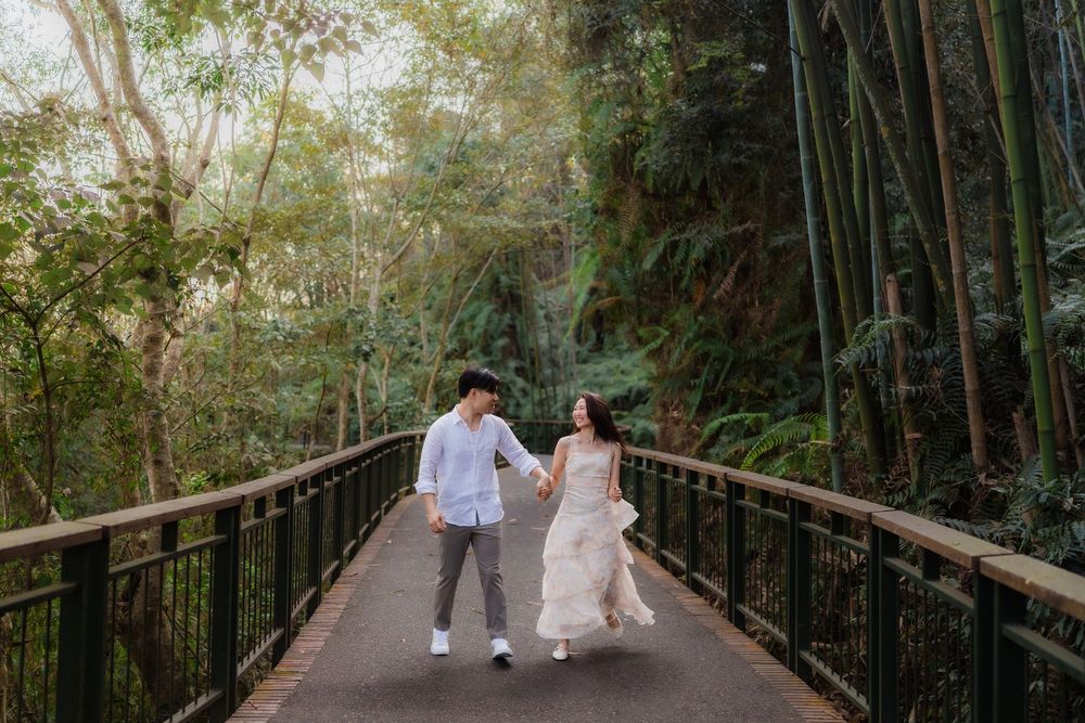 Couple holding hands and walking on a bridge surrounded by lush green bamboo forest in a romantic outdoor setting.