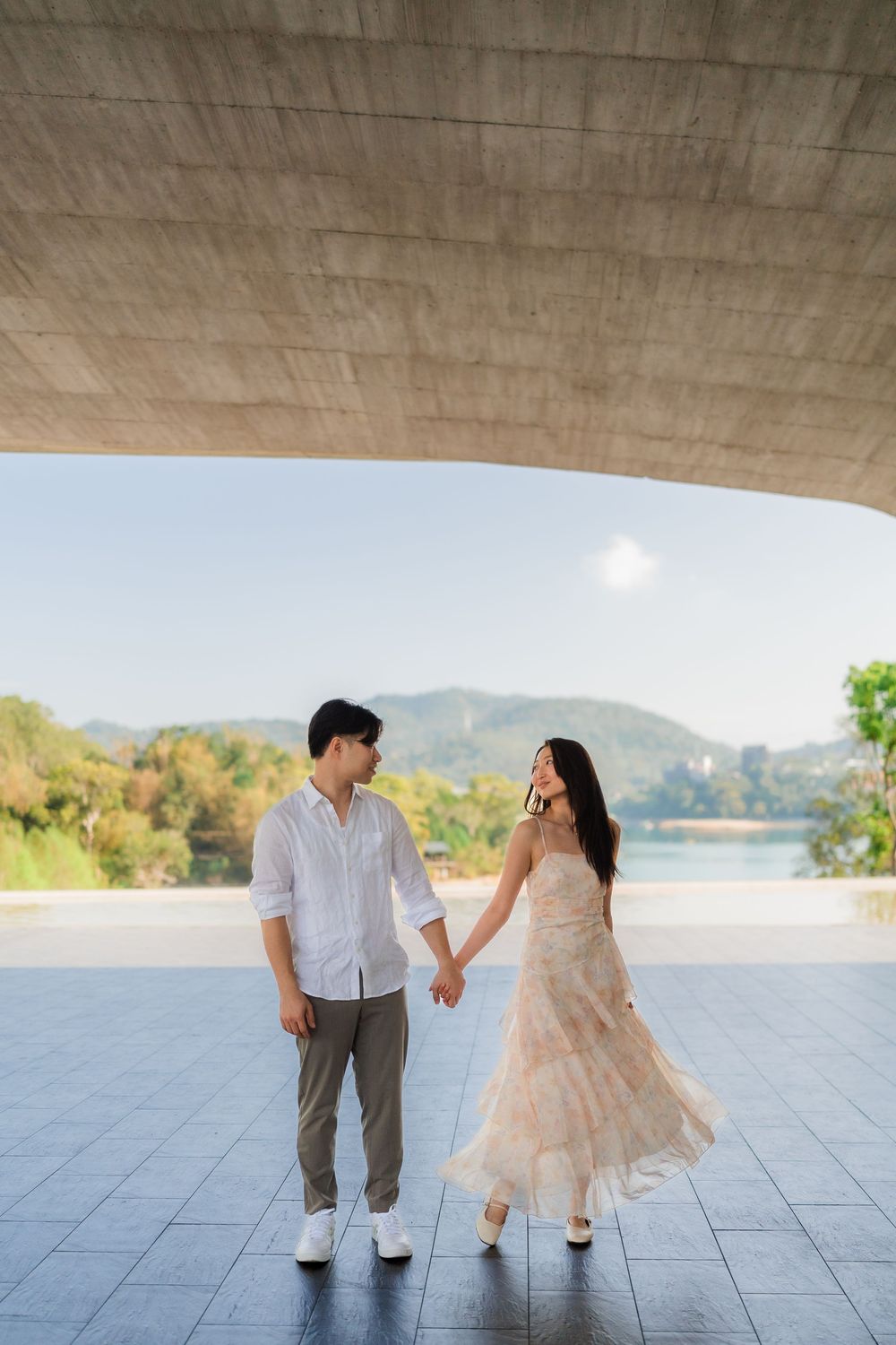 A couple holds hands and gazes at each other under a curved concrete structure with a scenic lake backdrop.