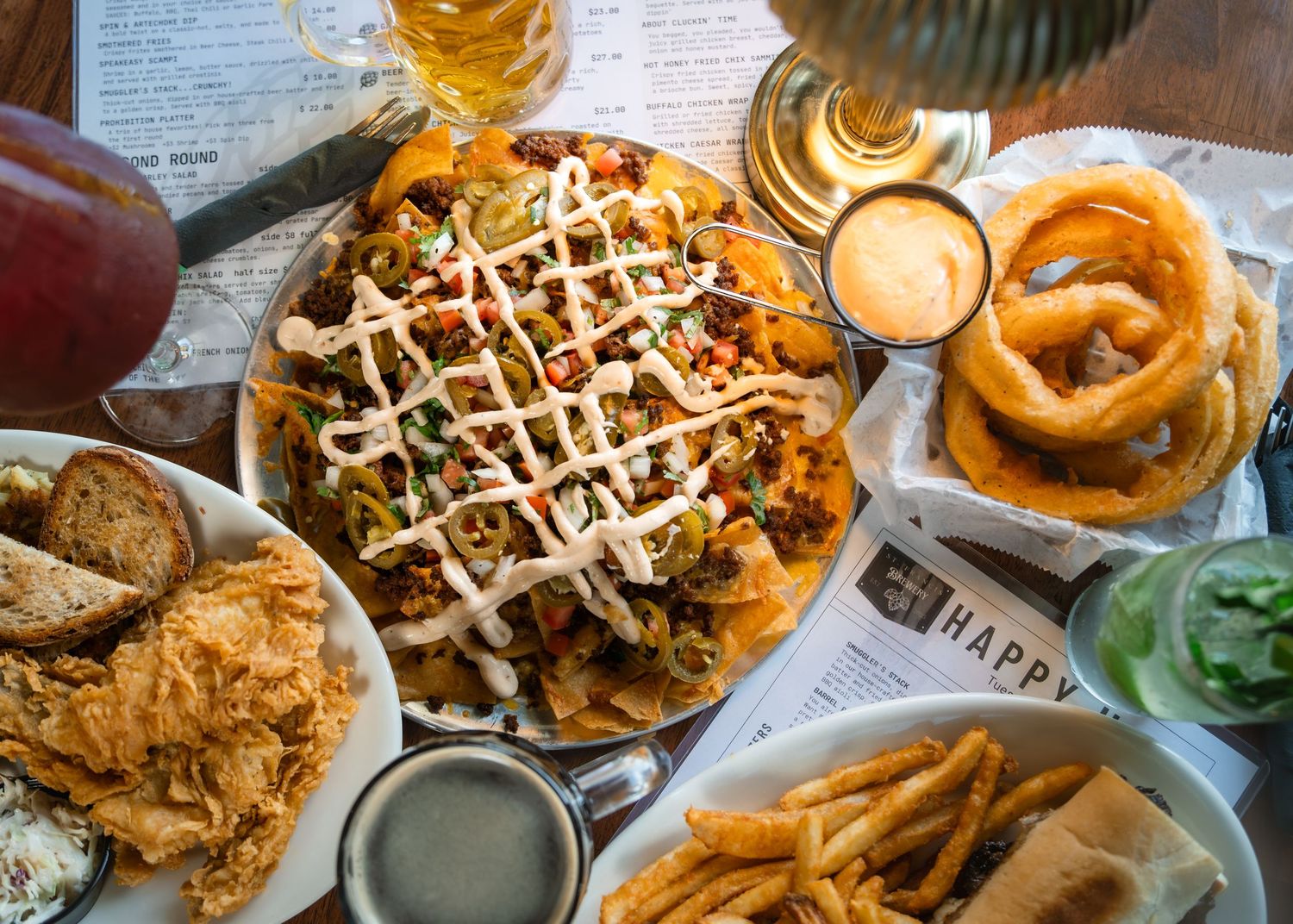 A spread of bar food including loaded nachos, onion rings, fries, and fried chicken on a restaurant table.