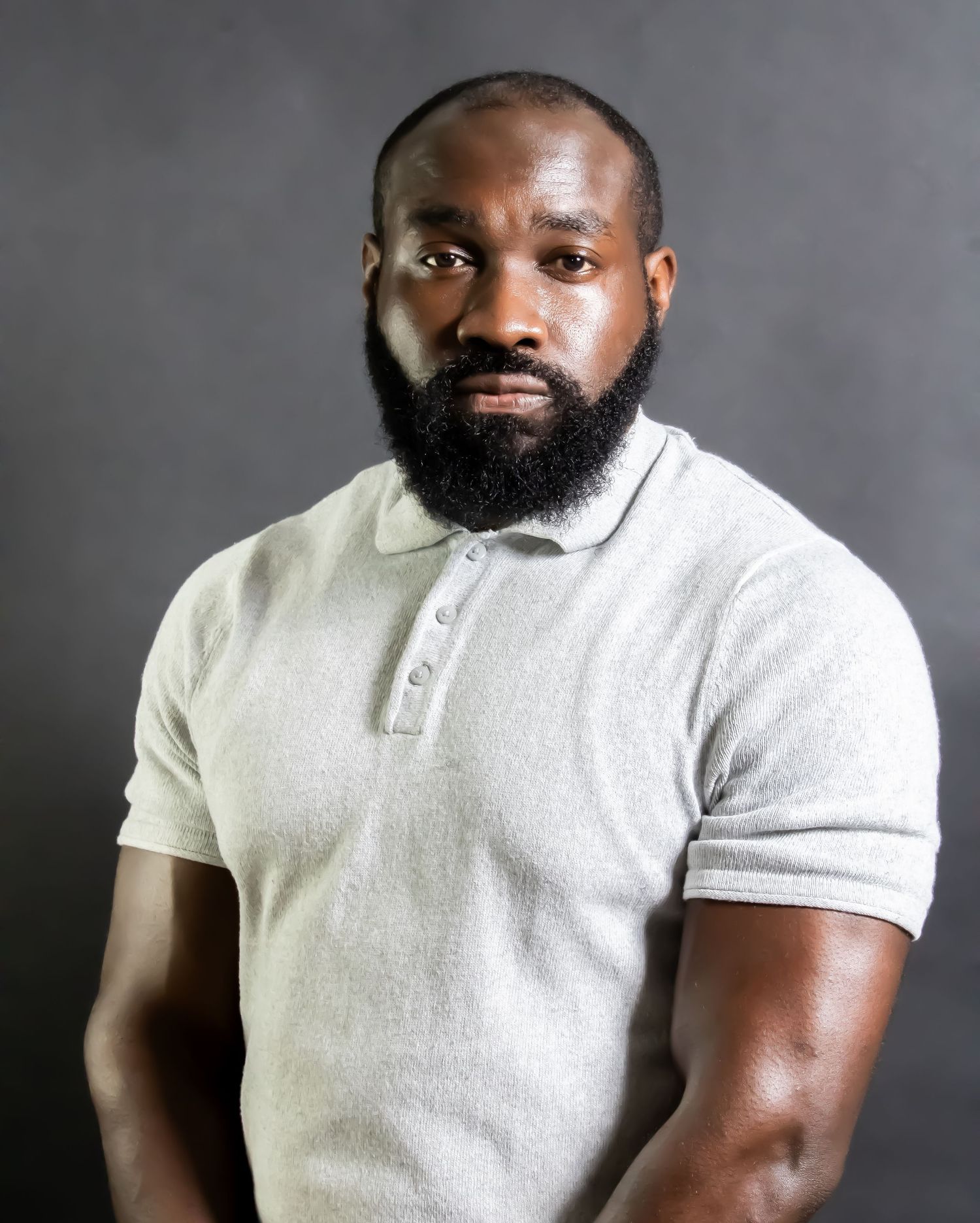 Muscular Black man with beard wearing gray polo shirt poses for professional headshot photo.
