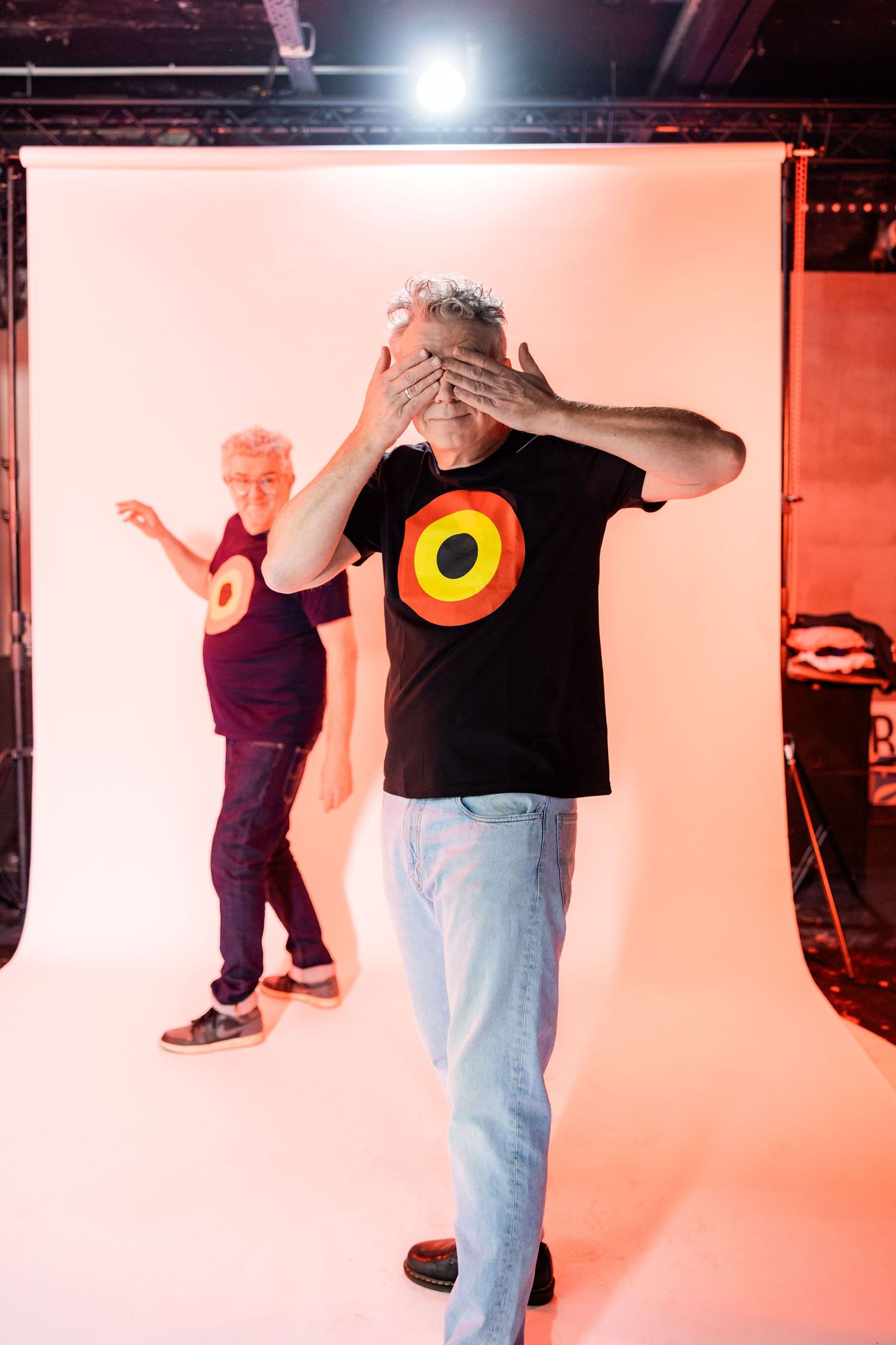 Two people in a photography studio, one in a black target t-shirt covering his eyes, another pointing behind him.