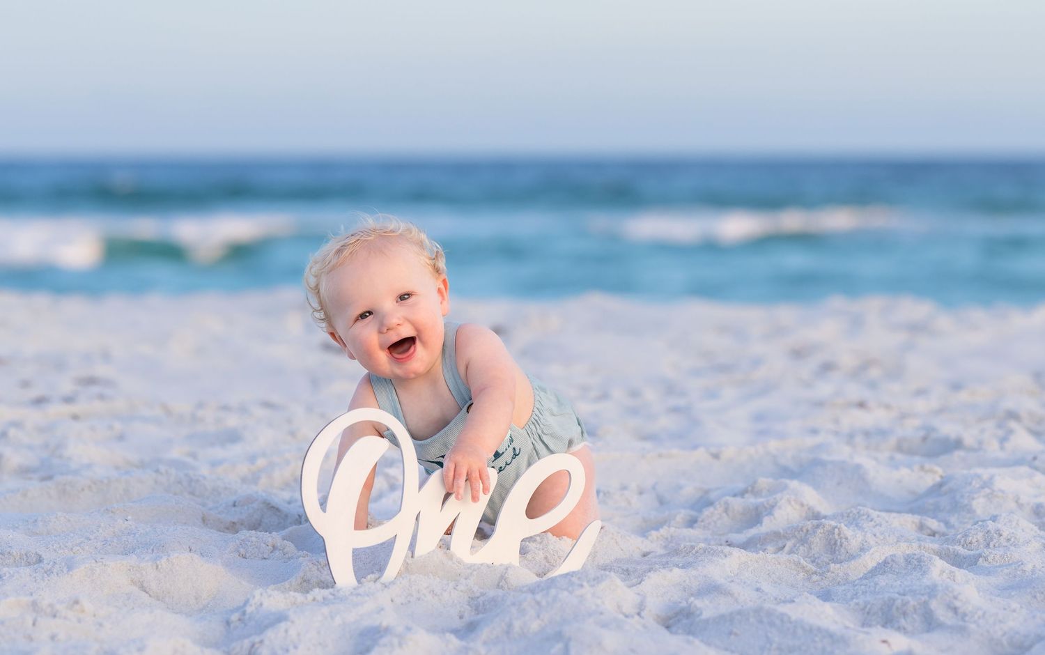 Happy smiling baby sitting on white sand beach next to a decorative One sign.