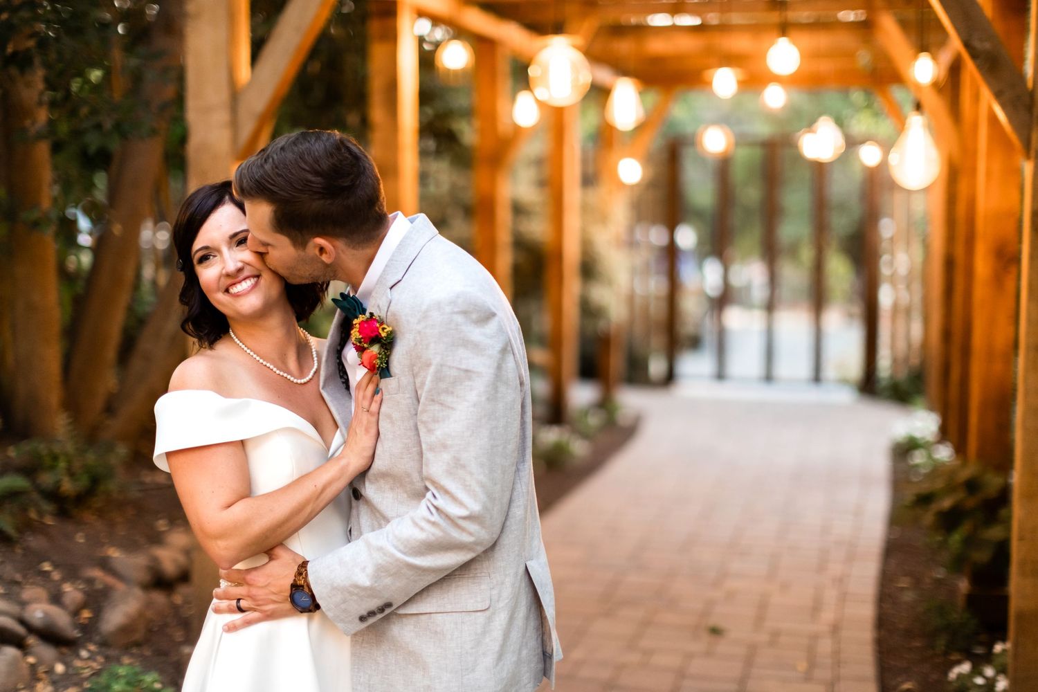 Bride smiling as groom kisses her cheek under wooden pergola with string lights at outdoor wedding venue.