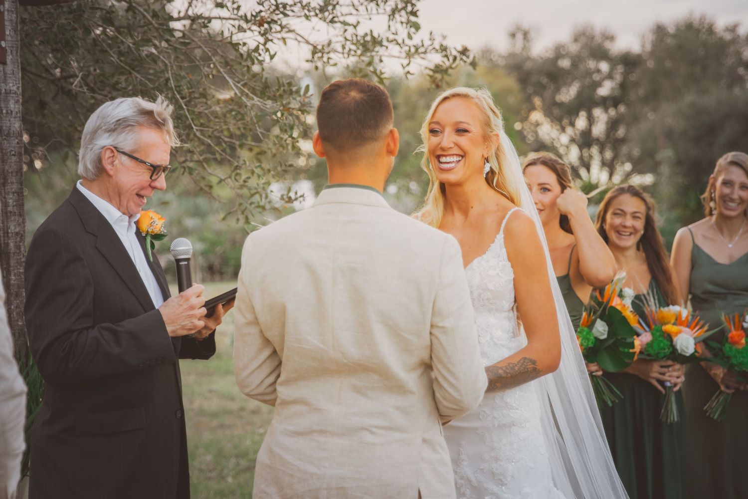 A bride laughs with joy as the groom says his vows to her during an outdoor wedding with green and orange hues