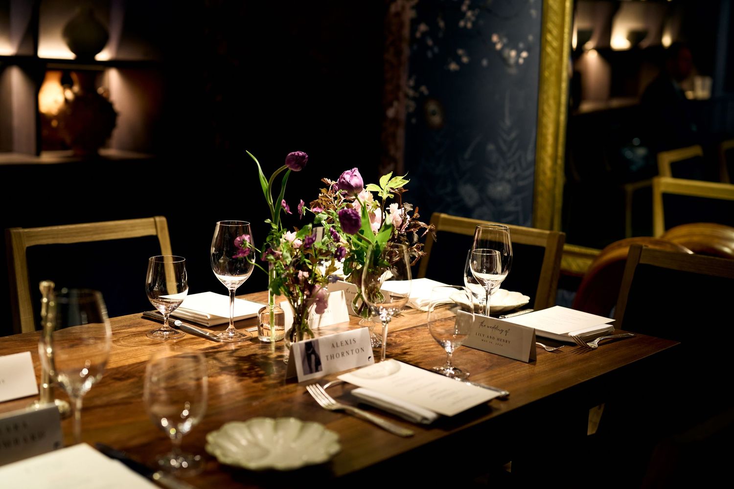 Elegant restaurant table set for dining with floral centerpiece, wine glasses, and white napkins in a dark, moody interior.
