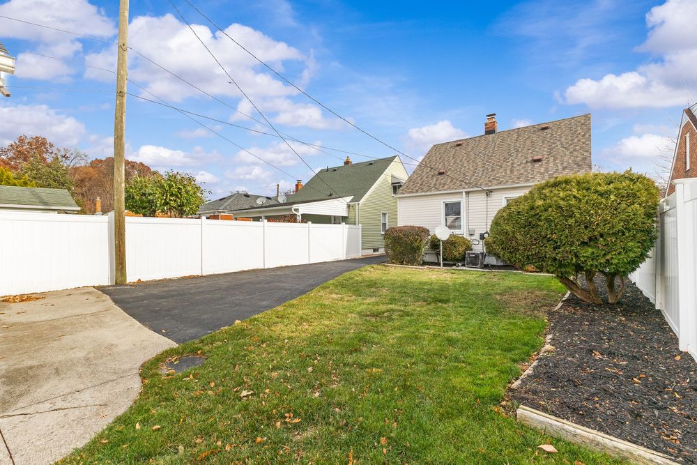 Backyard with lush green lawn, white vinyl fence, driveway, and neighboring houses under a bright blue sky.