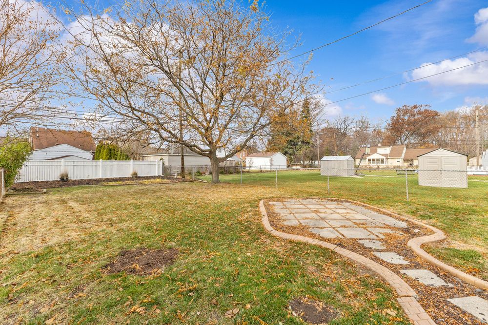 Spacious backyard with lush green lawn, large bare tree, stone pathway, white vinyl fence, and storage shed on a clear day.
