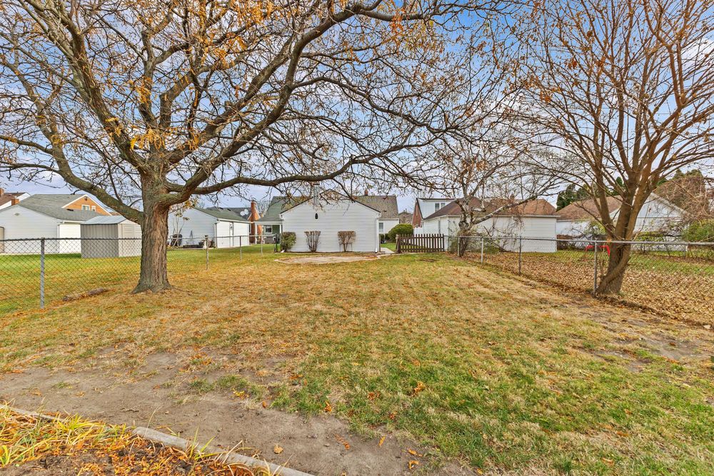 Spacious suburban backyard with mature trees, green lawn, and white houses visible in the background on a cloudy day.