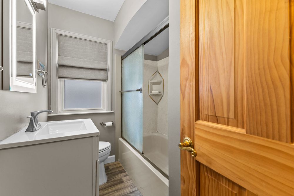Modern bathroom with sliding glass shower door, single vanity, toilet, and wood-grain flooring.