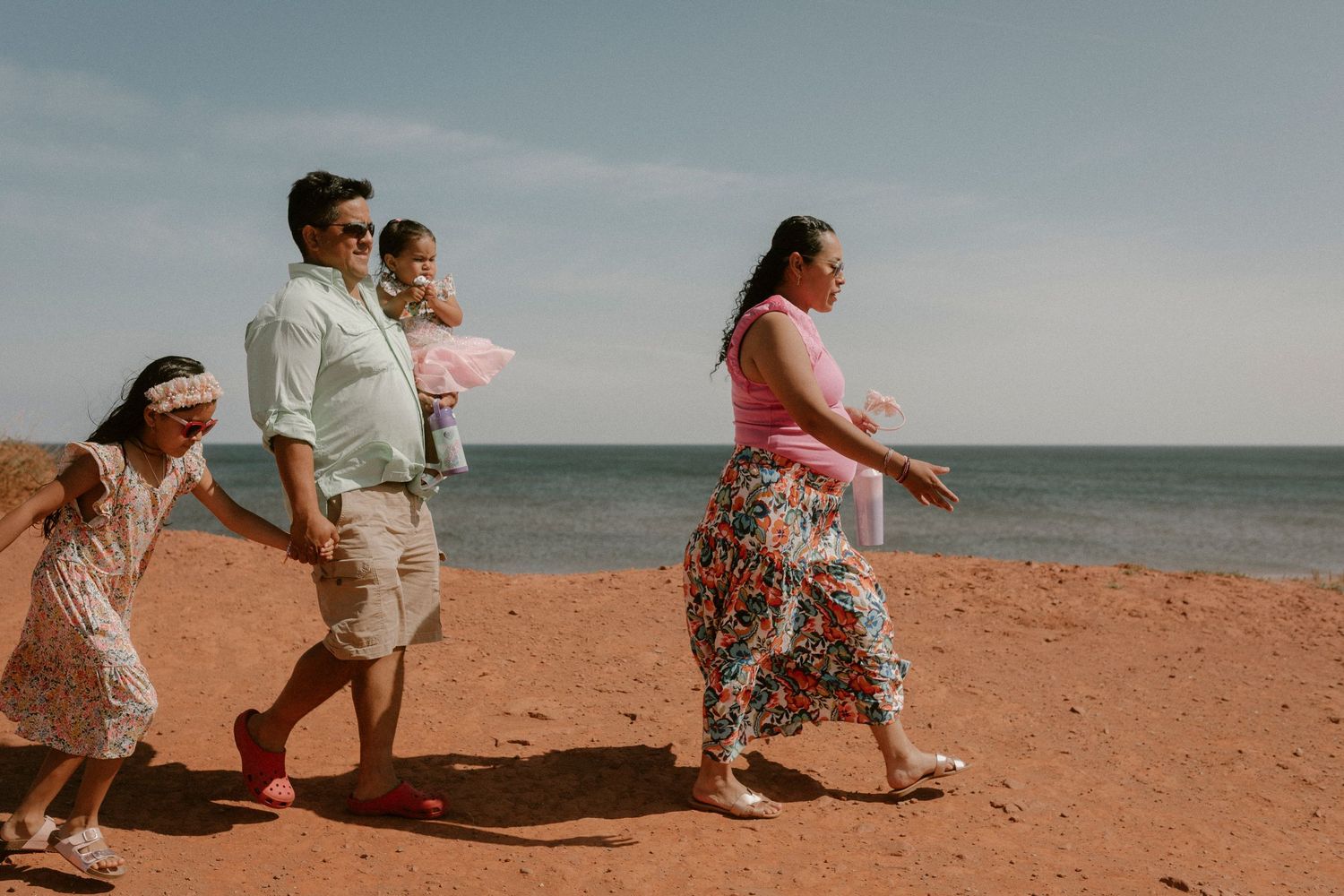 Family of four walking along a red sand beach on a sunny day by the ocean.