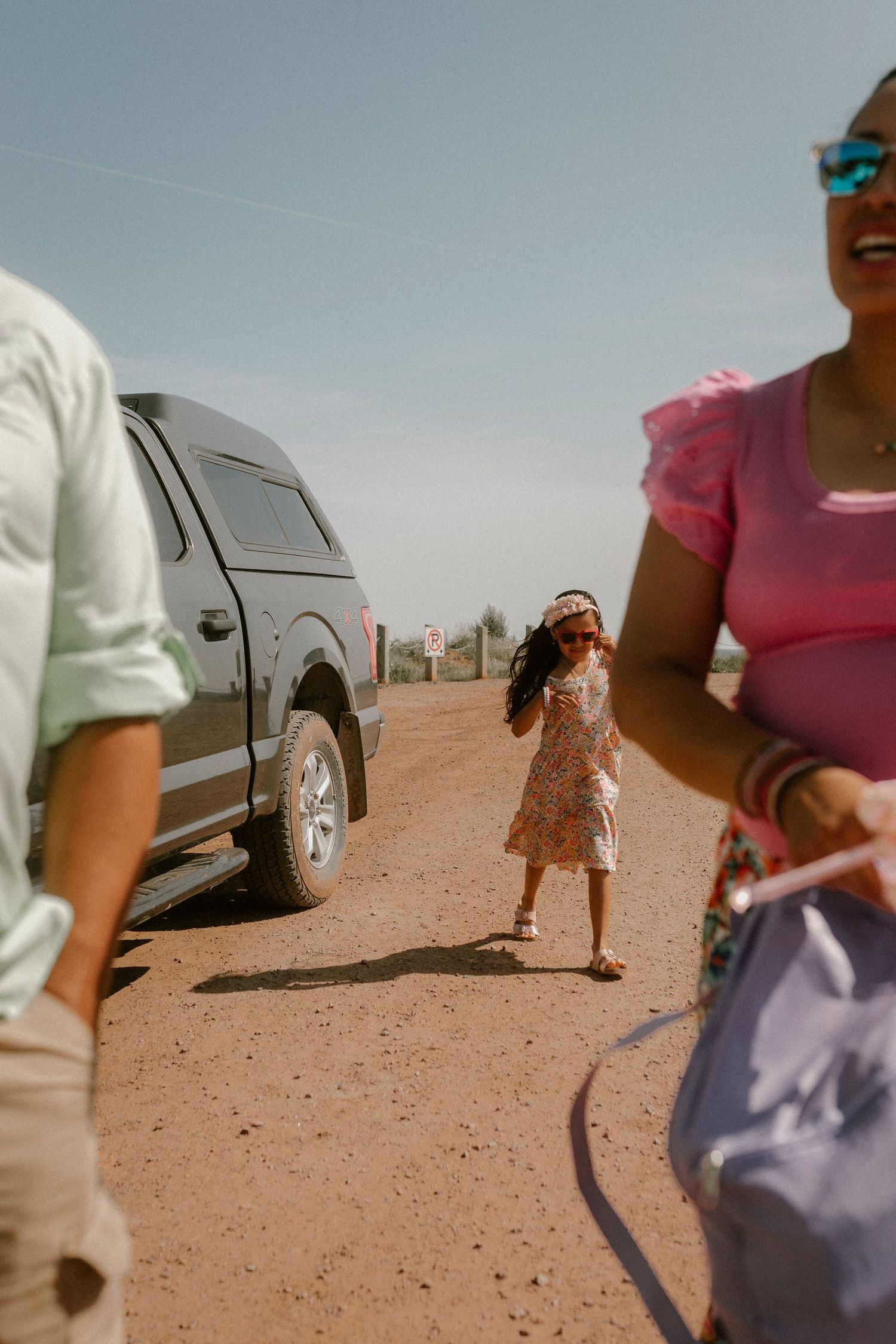 Young girl in floral dress standing near a truck on a dirt road on a sunny day.