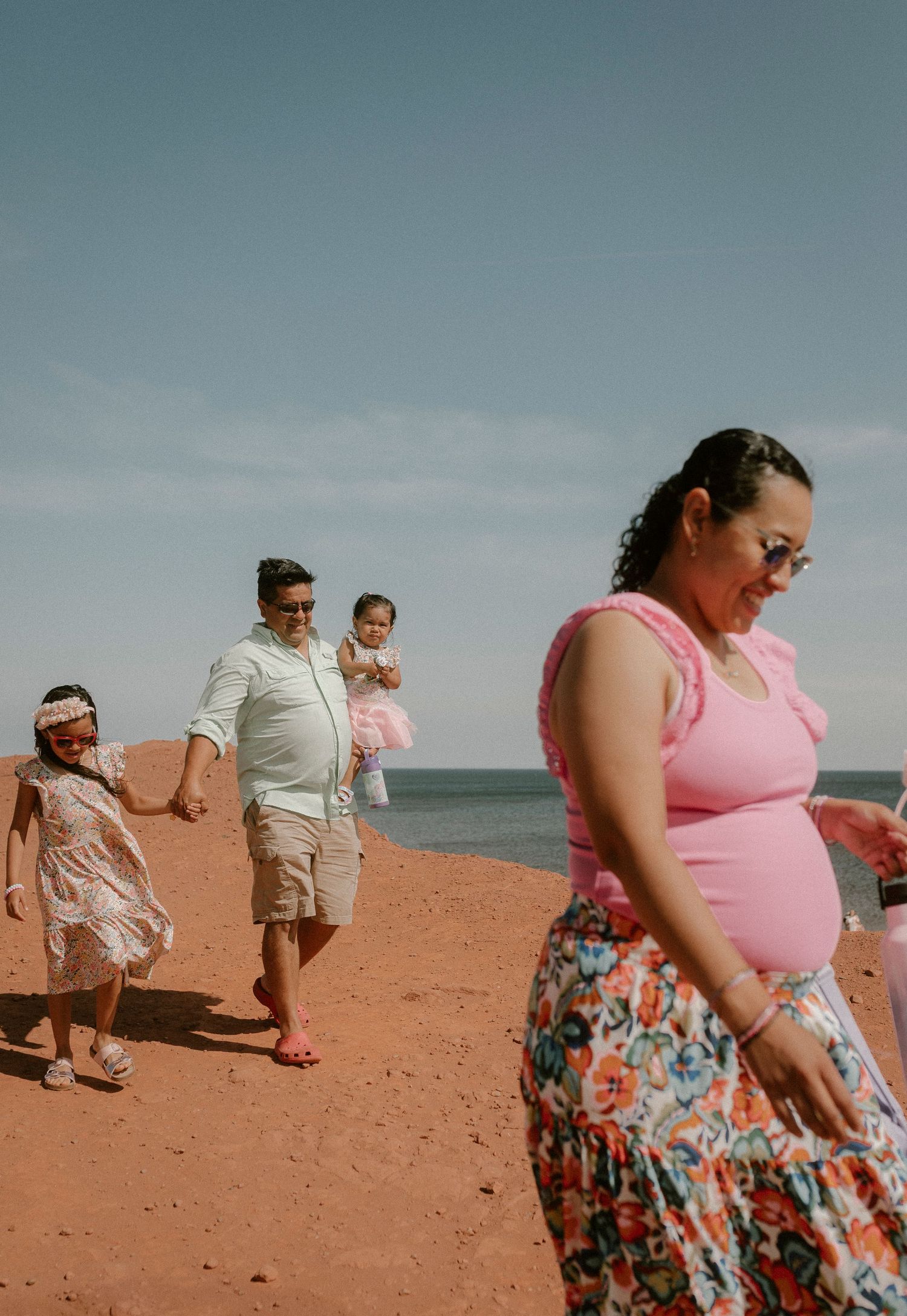 Pregnant woman in pink top walks with family including man carrying toddler on PEI red sand beach near ocean.