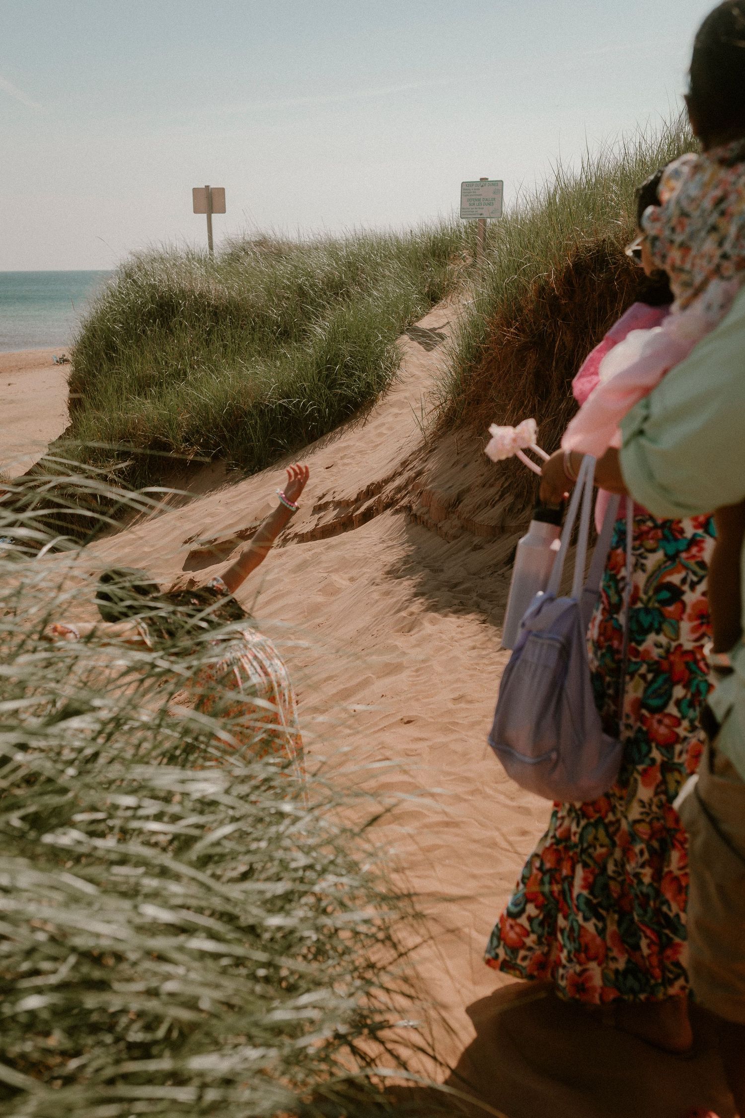 A woman in a floral skirt stands near sandy dunes with beach grass as a child reaches up from below.